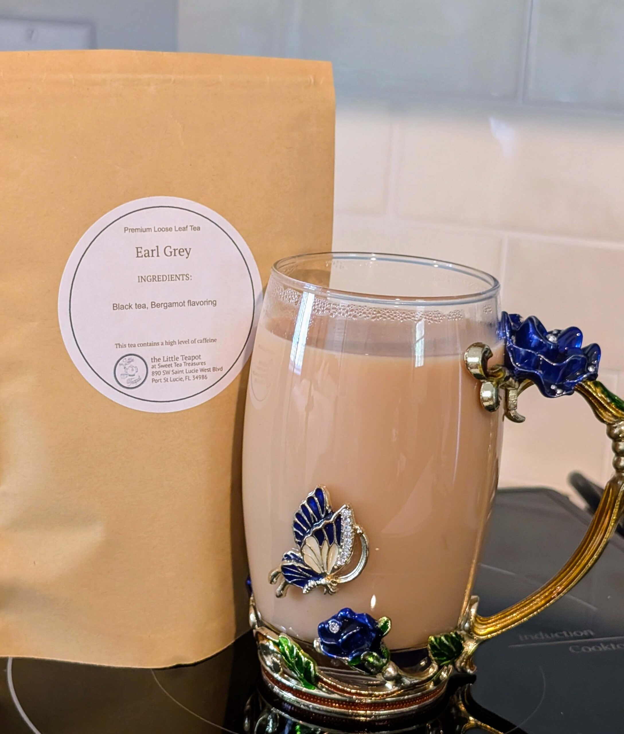 A packet of loose earl grey tea, next to a beautify clear glass tea mug with blue flowers, and a blue butterfly on it.