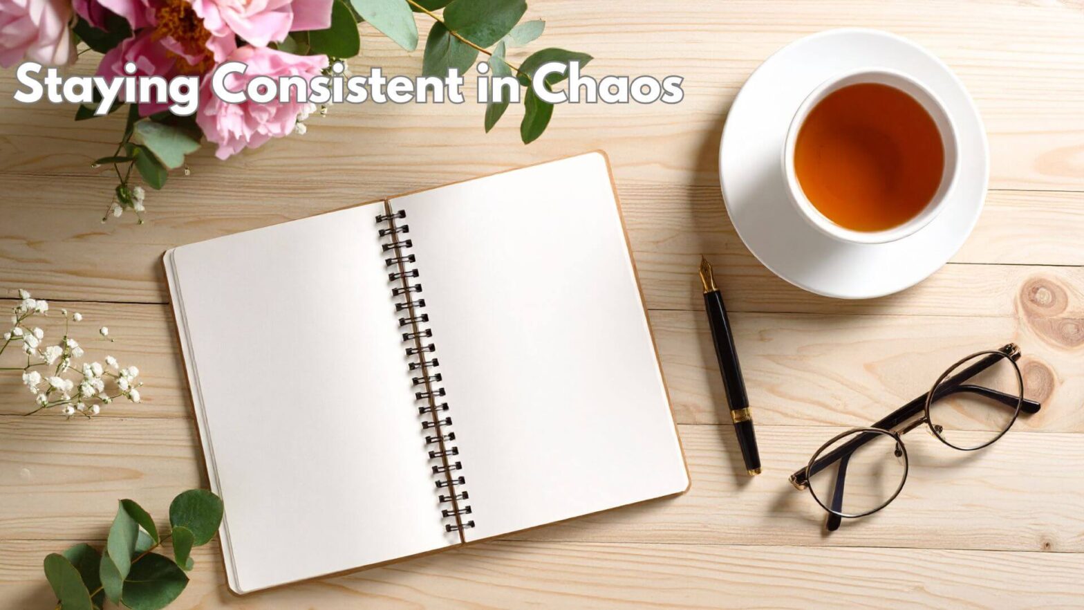A journal, cup of tea in a white cup and saucer, a pen, and a pair of glasses surrounding it. there are flowers in the left hand corner and text that says Staying Consistent in Chaos.