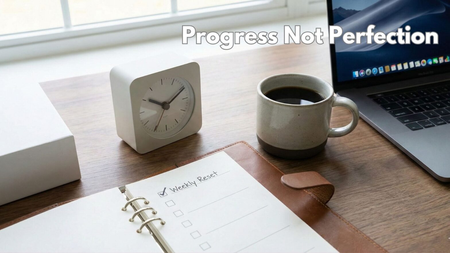 A picture of a wooden desk, with a desk clock, open calendar, coffee mug, and a laptop on it. With the words Progress not Perfection.