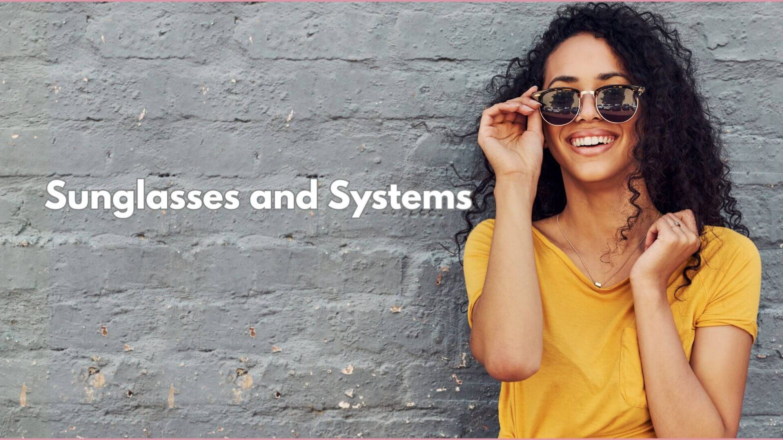 Woman with with dark curly hair and sunglasses wearing a yellow blouse, standing up against a grey painted brick wall. Text that reads Sunglasses and Systems.